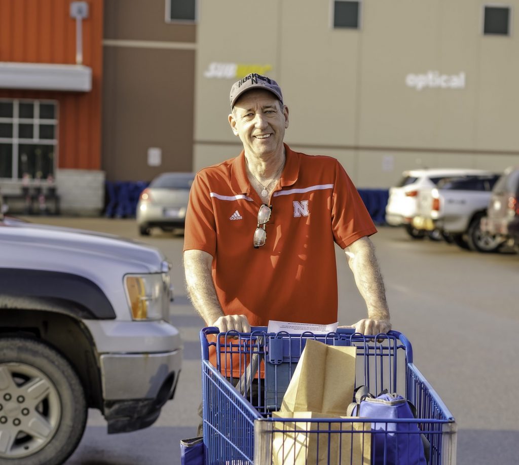 tall man with red shirt pushes grocery cart with groceries in the store parking lot