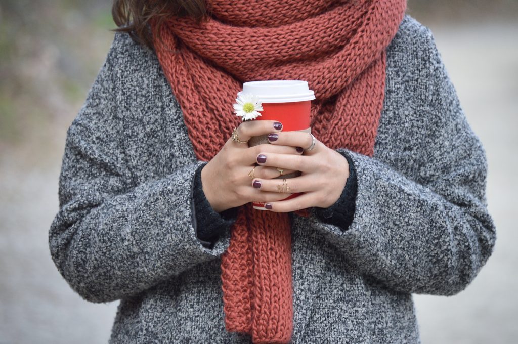 woman in winter coat and scarf holding red to go coffee cup with small daisy